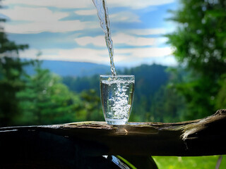 Aufkleber - Crystal clear water pours into a glass, sits on rustic wood, with a scenic mountain backdrop.  Freshness and nature are beautifully captured.