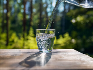 Aufkleber - Refreshing clear water pours into a glass on a wooden surface outdoors, sunlight illuminating the scene amidst a lush green forest backdrop.