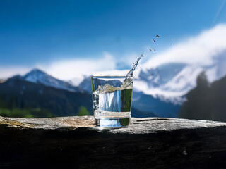 Aufkleber - A glass of fresh water sits on a rock, with a majestic mountain range and clear sky in the background. The water splashes dynamically, suggesting purity and refreshment.
