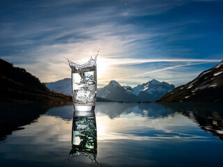 Aufkleber - A glass of ice water sits by a pristine mountain lake at sunrise, its reflection mirrored perfectly on the still surface.  The moment of splashing is captured, creating a vibrant scene.