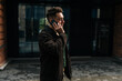 © dikushin - Side view of stylish young man making phone call standing outside modern office building, enjoying seamless communication in urban environment. Concept of modern successful city lifestyle.