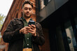 © dikushin - Low-angle view of handsome bearded man standing on city street, using smartphone to check messages, browse internet, stay connected enjoying urban environment. Concept of urban lifestyle.