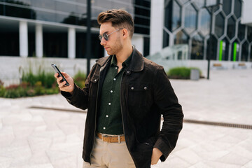 Wall Mural - Stylish businessman wearing sunglasses holding using mobile phone, browsing internet, reading message walking near modern office building on city street during daytime. Concept of urban lifestyle.