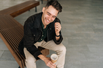 Wall Mural - Top view of cheerful man sitting on city bench, enjoying coffee break in vibrant urban setting, laughing while savoring moment and embracing simple pleasures of life. Concept of urban lifestyle.