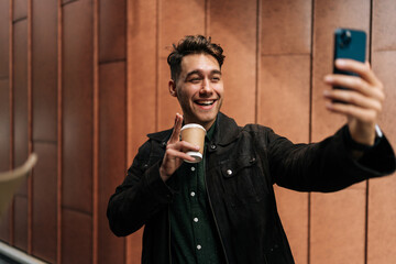 Wall Mural - Positive man making video call using smartphone holding coffee cup, standing in urban environment, looking to screen with happy expression. Concept of modern communication and urban lifestyle.