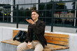 © dikushin - Successful businessman sitting on bench outdoors, holding takeaway coffee and recording voice message on smartphone, enjoying break time near office building. Concept of successful city lifestyle.