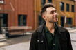 © dikushin - Portrait of handsome bearded young man wearing dark jacket and green shirt smiling looking away, standing in urban setting filled with contemporary architecture and cheerful atmosphere.