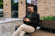© dikushin - Happy handsome young man sitting on bench with takeaway coffee and using smartphone smiling looking to screen on city street. Bearded male in casual clothes reading message on cellphone on autumn day