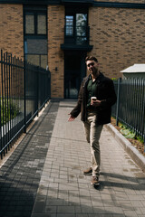 Wall Mural - Full length vertical portrait of handsome young man holding coffee cup standing by residential building with metal fence and brick walls, smiling looking at camera, enjoying peaceful moment outdoors.