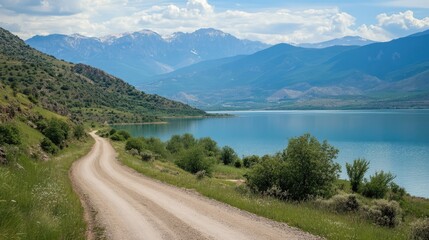 Wall Mural - winding dirt road along serene lake with majestic mountains under blue sky in a picturesque countrys