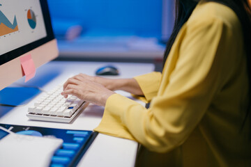 Wall Mural - Businesswoman wearing yellow jacket is working at night in office typing on keyboard and analyzing financial charts displayed on computer screen and tablet