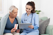 © amedeoemaja - Young friendly nurse or doctor smiles while explaining and showing an elderly woman patient the medications on a digital tablet. At home, in a medical clinic, or in hospice care. Concept of healthcare