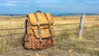 © Sirapopmillion - Migration Trends, Vintage leather backpack resting against a rustic fence in a golden grassy field under a bright blue sky with distant hills, symbolizing adventure and exploration