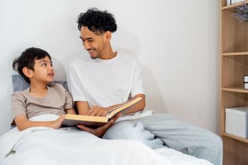 Asian young father sit near his son on bed and read the book also look each other with happiness.