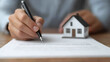 © praewpailyn - close up of hands signing property insurance document with model house nearby, symbolizing home ownership and security