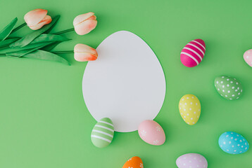 Spring composition with colorful Easter eggs and tulip flowers on pastel green background with paper card note copy space. Minimal Easter concept. Creative festive holiday flat lay. Easter aesthetic.