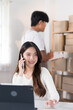© snowing12 - A woman uses her phone to take customer orders while working on her laptop, surrounded by boxes and clothing racks