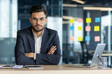 Sticker - Latin American businessman wearing glasses in an office sitting at a desk with arms crossed expressing confidence. Visible items include a laptop, and notes, indicating a professional environment.