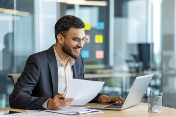 Wall Mural - Latin American businessman in a formal suit smiling while working on a laptop in a modern office, holding documents and sitting at a desk surrounded by professional workspace items.