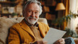 © Nataliia - Smiling senior man sitting comfortably at home, holding a letter and enjoying a cozy, warm atmosphere. Close-up