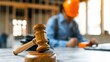 © Vilaysack - A gavel is in focus on a table, with a worker in a hard hat in the background, symbolizing law and construction.