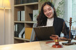 © Phimwilai - Asian lawyer smiling and reviewing legal documents on a clipboard, with a gavel and scales of justice on her desk, representing the legal profession and justice system