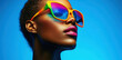 © Ramus - A woman confidently poses while wearing oversized, brightly colored sunglasses. Her striking features are illuminated by a vivid blue background, enhancing her vibrant style.