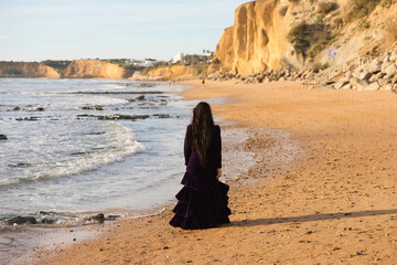 Wall Mural - beautiful young woman dancing flamenco walks along the shore of the beach at sunset, wearing a purple velvet dress with ruffles.