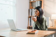 © kenchiro168 - young woman sits at wooden desk, holding cup while looking at laptop screen in cozy home office with bookshelves and soft lighting