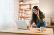 © kenchiro168 - cheerful woman multitasking while talking on phone and working on laptop at wooden desk in bright home office with shelves and decor in background