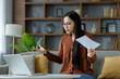 © Liubomir - Asian woman in home office setting, engaged in video call holding paper, discussing project. Laptop on table, remote work environment. Emblematic of modern communication and indoor workspace.