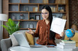 © Liubomir - Asian woman conducting online lesson from home. She sits on sofa with laptop, holding a whiteboard marked Lesson, engaged in interactive teaching session, conveying education and technology.