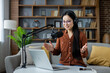 © Liubomir - Asian woman hosting a podcast at home, speaking into a microphone while using a laptop. Wearing headphones, she appears engaged and lively, surrounded by a comfortable setting. Smartphone visible.