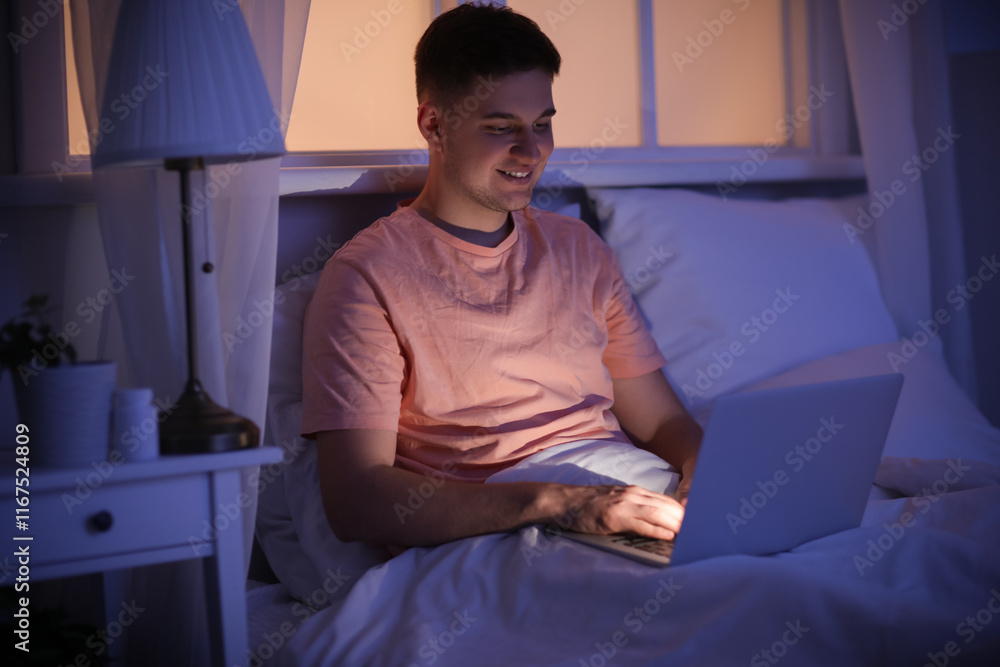 Handsome young happy man with laptop sitting in bed at night