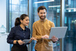 © Liubomir - Two colleagues man and woman inside office standing by window talking, group of office workers using laptop discussing new work project.