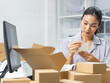 © kan - Portrait of Asian woman entrepreneur preparing a product for delivery, attaching a sticker shipping label to parcel box at warehouse office. Asian woman shop owner managing package for ship to client.
