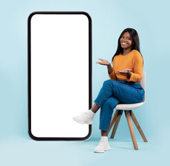 Full length of pretty black lady sitting on chair next to big blank smartphone with mockup for mobile app or website design, blue studio background, showing copy space for your online advertisement