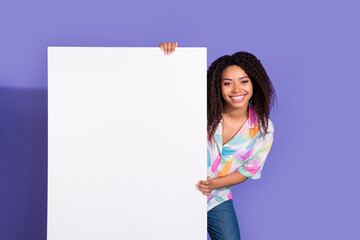 Smiling young woman with black curls standing against a vibrant purple background, holding a blank white sign