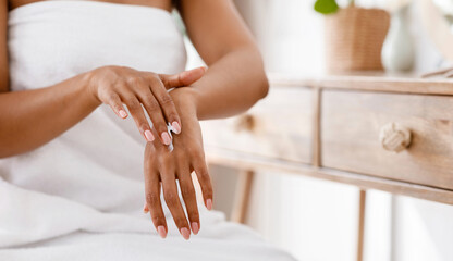 Wall Mural - Skin nutrition concept. Unrecognizable black woman applying moisturizing cream to her hands, sitting wrapped in towel in bedroom, cropped image