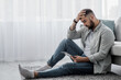 © Prostock-studio - Man is chatting on phone at home feeling stress, bad news, boredom alone. Sad despair serious millennial attractive bearded guy sit on floor, look at tablet in living room interior, copy space