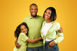 © Prostock-studio - Family portrait. Happy african american parents and cute daughter embracing and smiling at camera over yellow background. Excited black mother, father and child posing together in studio