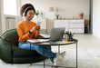 © Prostock-studio - People And Technology. Smiling young black lady sitting at tea table with laptop, holding using cell phone. Cheerful female in wireless headset watching video, chatting taking break, free copy space