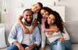 © Prostock-studio - Happy Loving Family. Portrait of cheerful African American man and woman sitting on the couch at home, posing for photo, smiling boy and girl embracing parents from the back. Bonding