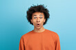 © Prostock-studio - A young African American man displays an expression of surprise with wide eyes and an open mouth, wearing an orange sweater, set against a bright blue backdrop.