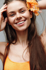 Poster - Cheerful young woman with long hair and orange flower, wearing bright orange top, smiling joyfully against a soft grey background.