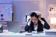 © crizzystudio - Young stressed businesswoman holding documents on head while working with laptop and financial reports in office at night, experiencing burnout and frustration from overwork