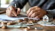 © Chanwit - A person is writing on documents with coins and bills scattered, alongside a small model house, symbolizing financial planning or real estate investment.