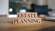 © E - Two wooden blocks stacked on a wooden table display 'ESTATE' and 'PLANNING' in dark serif font. A blurred indoor background creates a clean, professional focus on estate planning.