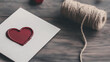 © alexandra_pp - A close-up of a handmade Valentine’s card featuring a deep red heart-shaped wax seal on textured paper, accompanied by a rustic twine spool, set on a wooden surface with warm, romantic tones.