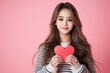 © Tondone - Smiling woman holding a red heart shape against a pink background, portraying love and happiness. The image conveys warmth and positivity in a simple and charming way.